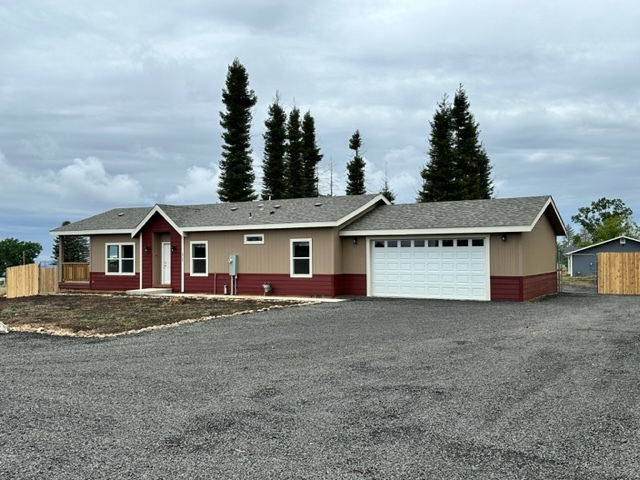 A photo of the exterior of a home in Paradise, California. There is a gravel driveway that leads to a 2-car garage. The home has dark red siding at the bottom and stopping at the base of the window and then tan siding from there to the top. There are evergreen trees behind the home and the sky is cloudy.