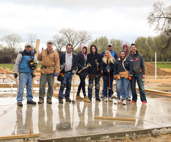 A photo of a group of homeowners standing on top of a concrete foundation for a new home. The concrete is wet from rain. The group is posing and smiling.