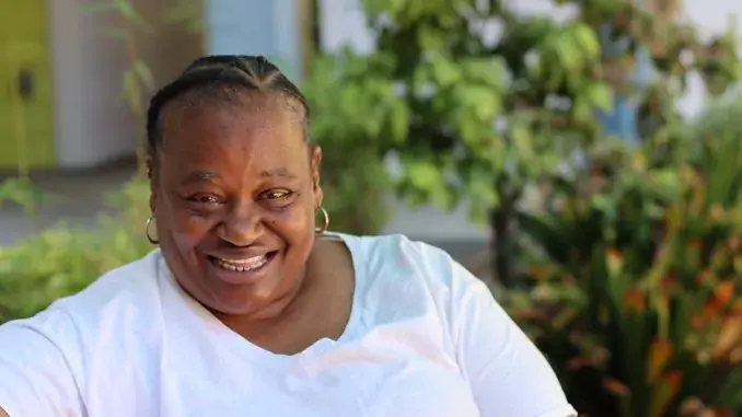 A resident at Maple Park Apartments sits in the outdoor courtyard. She is wearing a white tee shirt. She has black hair pulled back into braids. She has hoop earings. She is smiling.