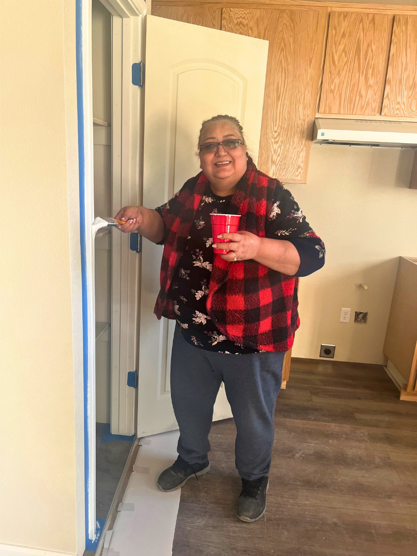 A homeowner is painting the trim around a pantry door in her kitchen. She is holding a paintbrush and a red cup that contains white paint. She is smiling.