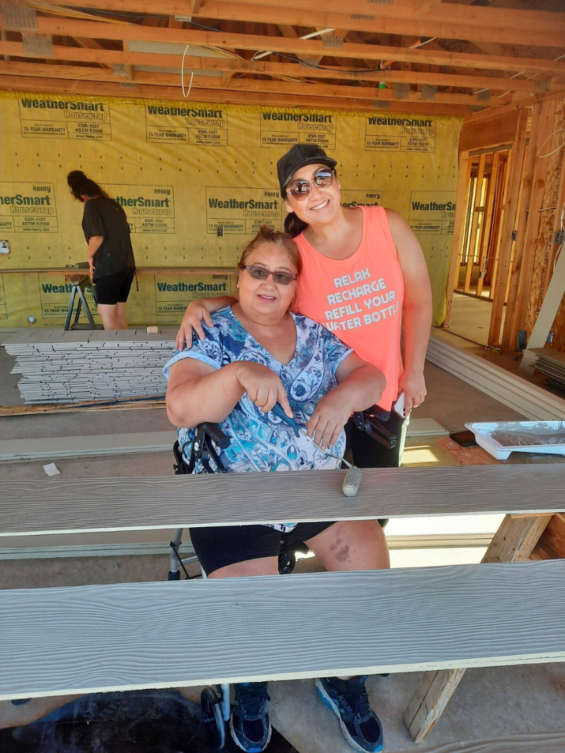 A homeowner sits and one of her daughters stands next to her. The daughter has her arm around her mother. The are in the garage. There is exterior siding in front them that is being prepared for painting. The homeowner and daughter are posing and smiling together.