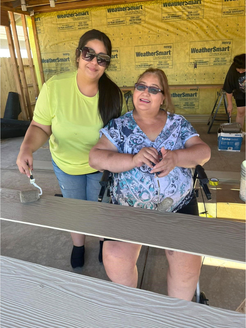 A homeowner and her daughter are in the garage. The homeowner is sitting and her daughter stands next to her and paints the wall siding that will be installed later. They are smiling.