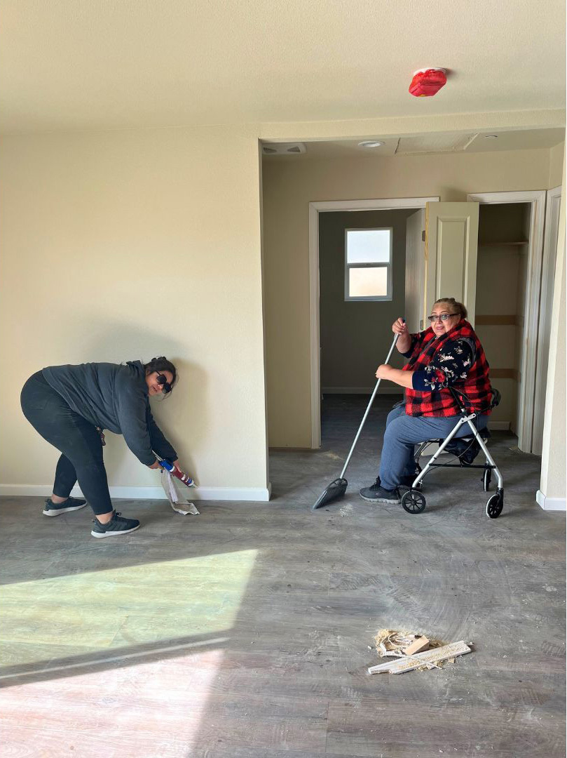 Two homeowners are working inside a home. The photo shows the living room with new flooring installed. The flooring is dusty from construction. One homeowner is bending over and applying caulk to the floorboards. The other homeowner is sitting down and using a broom to sweep off the dusty floors.