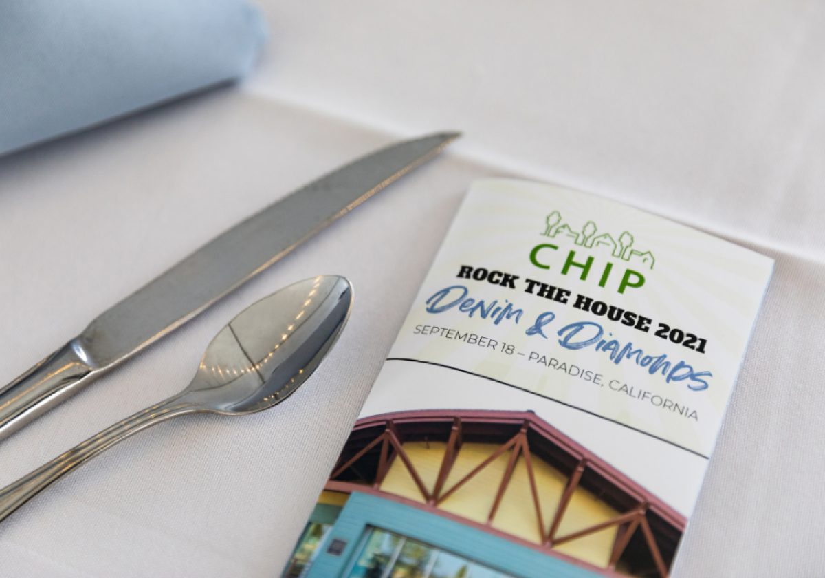 A photo of a place setting consisting of a silver knife and spoon on a white linen table and a program rests on the table as well. The program is for the fundraiser from 2021. It shows the CHIP logo and reads "Rock the House 2021 Denim and Diamonds."
