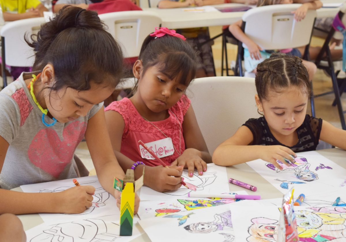 A photo of 3 young girls sitting at a table doing an art project. On the table are multiple coloring sheets, a box of crayons, and additional crayons scattered around. The girls are focused on their coloring.