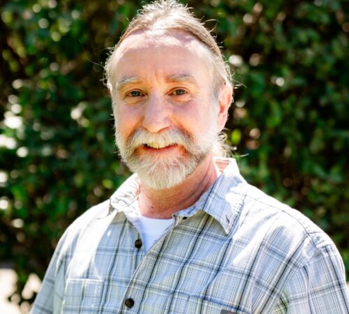 This is a portrait photo of Jason Marr, a staff member. He is wearing a gray and white plaid shirt. He has brown hair pulled back and a gray beard. He has a slight smile.