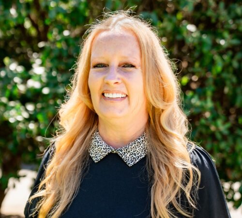 This is a portrait photo of Holly McCormick, a staff member. She is wearing a black shirt with a white leopard print collar. She has long blonde hear past her shoulders. She is smiling.
