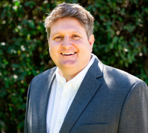 This is a portrait photo of Mark Montgomery, a staff member. He is wearing a white collared shirt and a gray jacket. He has short white and brown hair and is smiling.
