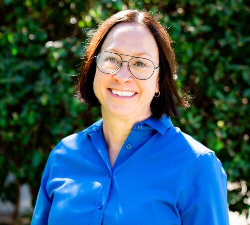 This is a portrait of Seana O'Shaughnessy, the President and CEO. She is wearing a blue top with long sleeves. She has short brown hair and is wearing glasses. She is smiling.