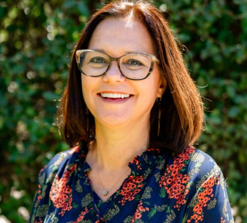 This is a portrait photo of Wendy Philips, a staff member. She is wearing a blue top with an orange-red flower pattern and green leaves. She has shoulder-length dark brown hair. She is smiling and is wearing brown glasses.