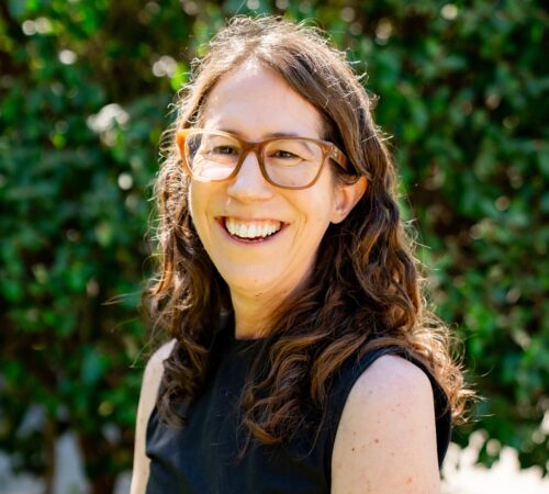 This is a portrait photo of Jill Quezada, a staff member. She is wearing a black top. She has brown curly hair that goes just beyond her shoulders. She is wearing glasses. She is smiling.