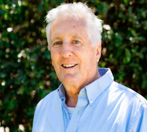 This is a portrait photo of Andy Holcombe, a board member. He is wearing a blue dress shirt. He has short white hair. He is smiling.