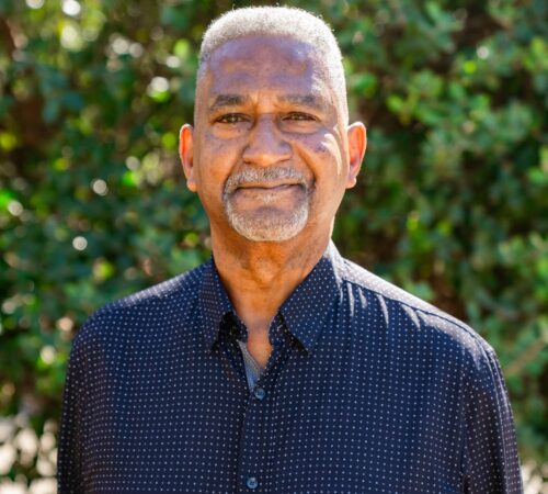 This is a portrait photo of David Goodson, a board member. He is wearing a blue-ish purple dress shirt with a small white polka dot pattern. He has a mix of short, dark, and gray hair and a goatee. He has a slight smile.