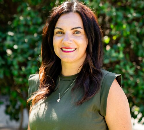 This is a portrait photo of Vanessa Haro, a board member. She has dark brown hair that is wavy and goes past her shoulders. She is wearing a black top. She is smiling.