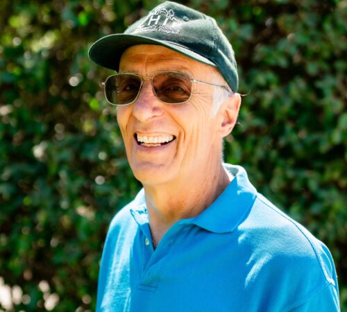 This is a portrait photo of Mark Hooper, a board member. He is wearing a light blue dress shirt and a green hat with the CHIP logo. Mark has white hair and wears glasses with transitional lenses. He is smiling.