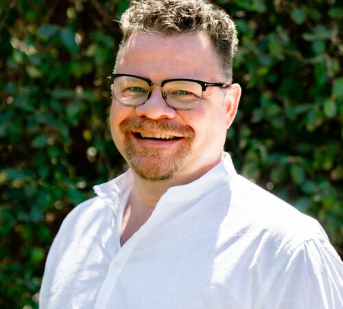 A portrait photo of Earl Jessee, a board member. Earl is wearing a white collared shirt. He has short, brown hair and a goatee. He is wearing glasses and has a slight smile.