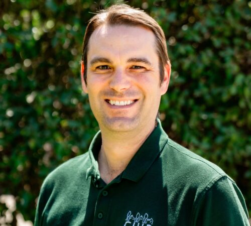 A portrait photo of Ross Simmons, a board member. Ross is wearing a green shirt with a white CHIP logo on the upper right side. He has brown hair and is smiling.