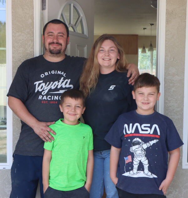 A family stands in front of the front door of their new home. A husband and wife stand in the back and two young boys stand in the front. The are posing and smiling.