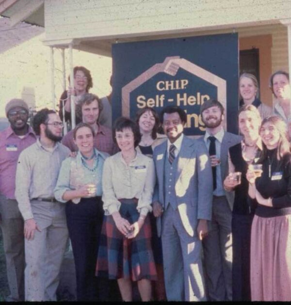This is a photo taken in the 1970s that shows the CHIP staff standing in front of a home that acted as its office at that point in time. The staff are posing for the picture and smiling. Many of them are wearing formal attire. The sign the hangs from the front porch of the office reads "CHIP Self-Help Housing."