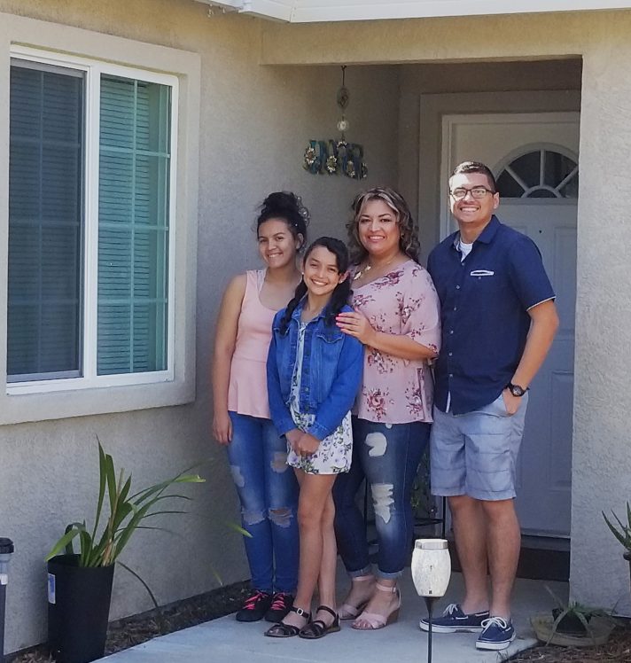 A family standing in front of the front door of their recently built home. A husband and wife stand next to their two daughters. They are smiling.