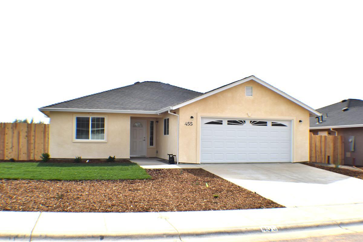 A photo of the exterior of a newly constructed single-family home. The photo is taken from the vantage point of the street and the view is of the front of the home and its front yard. The house is yellow, there is a garage and a window in the front of the home. The yard has a small area of grass and then bark covers the area between the sidewalk and the middle of the yard with the lawn begins.