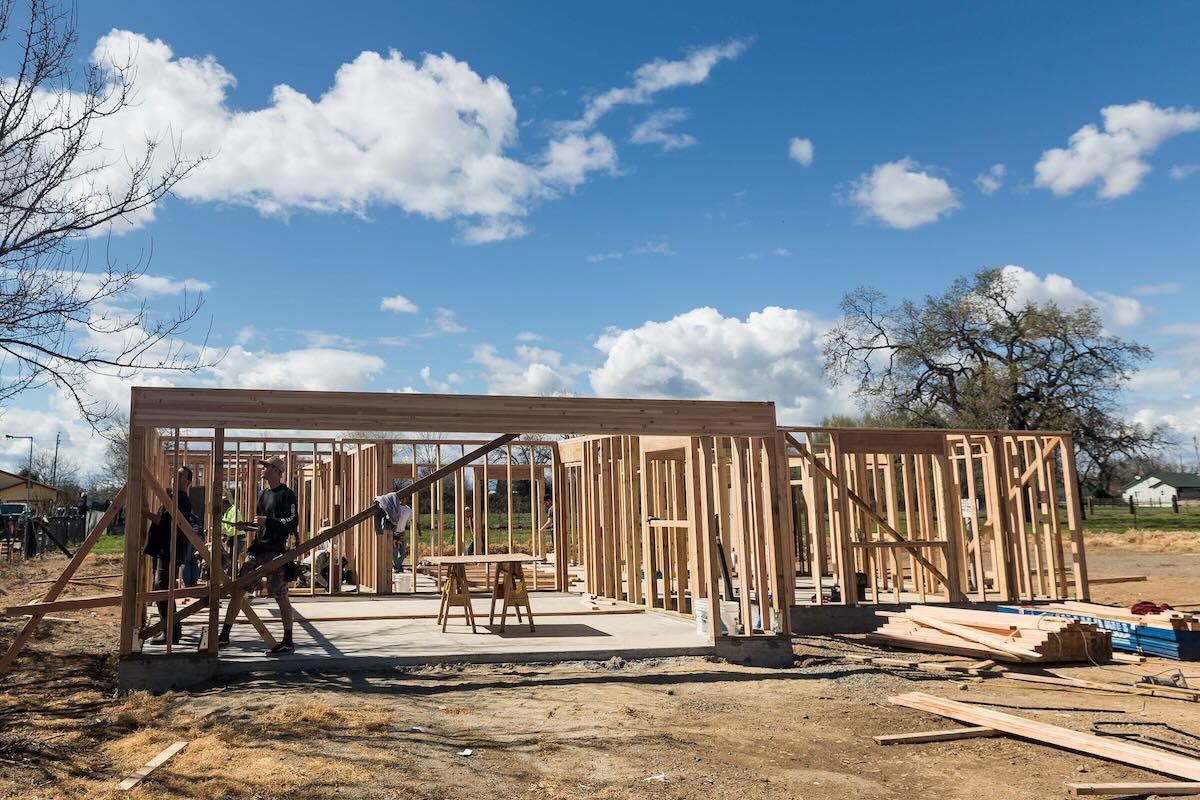 A photo show a single-family home under construction. The home is framed with two-by-fours but the roof has not yet been framed. In the background, there are some trees and a cloudy day.