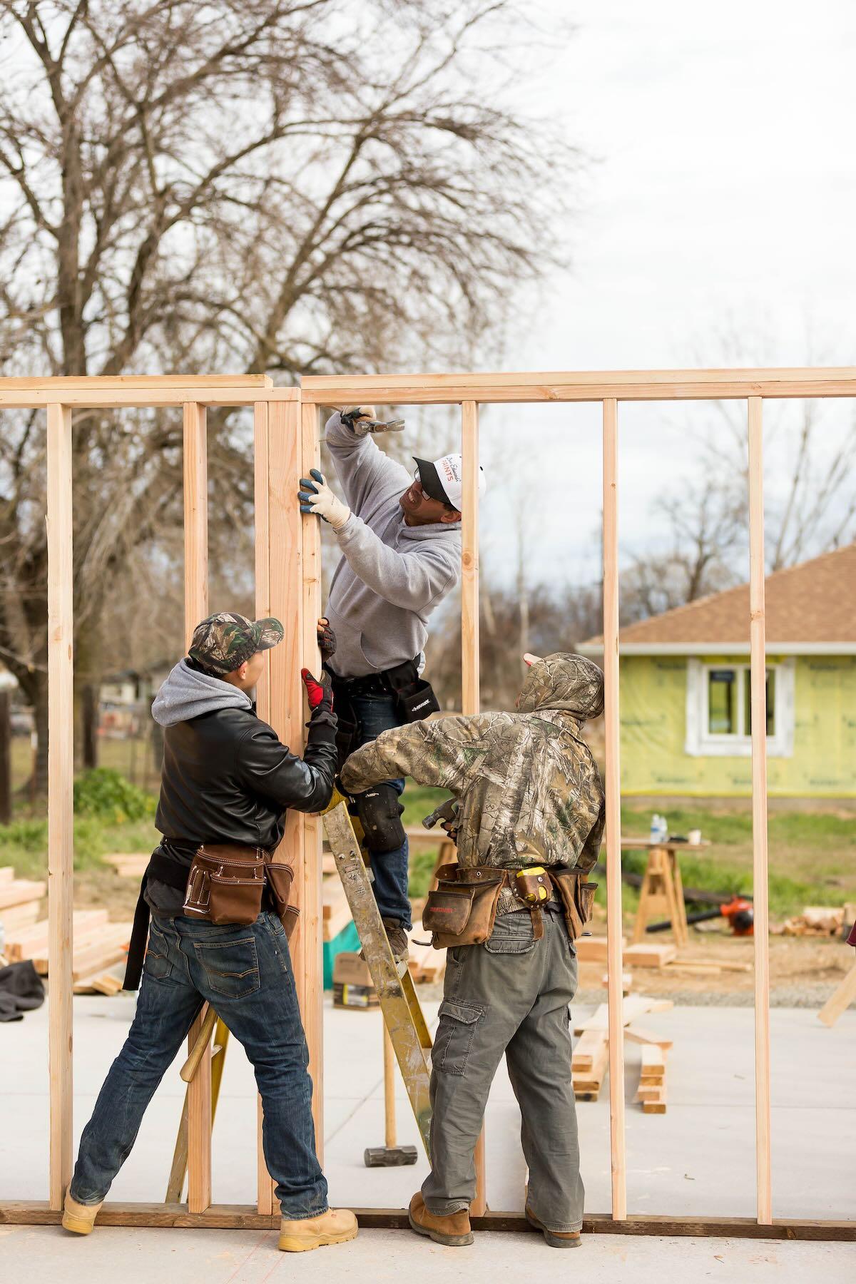 A photo shows a team of volunteers working on a single-family home.  They are working on a wall made of two-by-fours. One of them is standing on a ladder and hammering a nail.