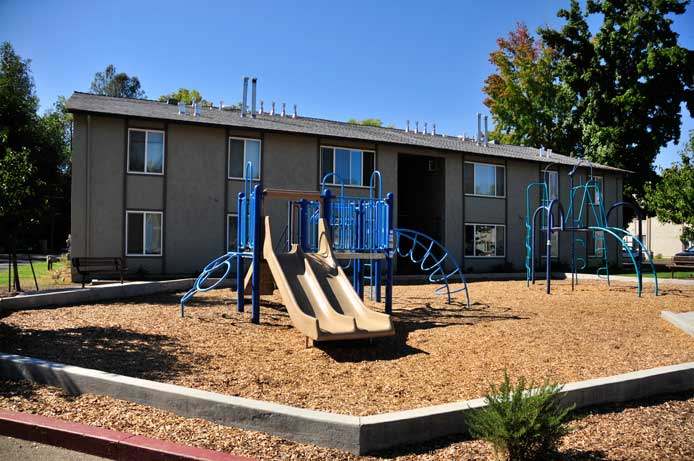 A photo of the playground at Brickyard Creek Apartments. It shows a brow plastic side-by-side slide and blue bars and green bars. The ground consists of playground bark. In the background is one of the buildings of Brickyard Creek Apartments and a few trees.
