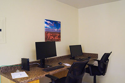 A photo of the computer lab shows two computers on a counter. Two computer chairs are tucked into the counter next to the computers. The background is a white wall with an art poster.