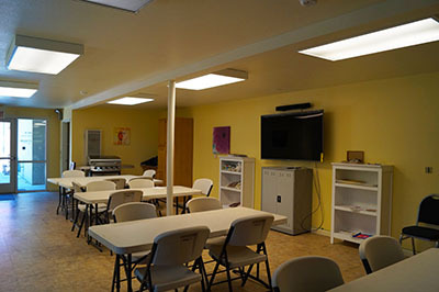A photo of the community room at Brickyard Creek Apartments. The room has 4 tables each with 4 chairs. The walls are painted yellow and one wall has a large flat-screen TV.