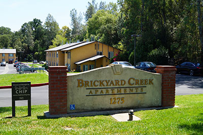 A photo showing the sign at the entrance of Brickyard Creek Apartments. The sign reads "Brickyard Creek Apartments" and in the background, you can see one of the buildings in the apartment complex.