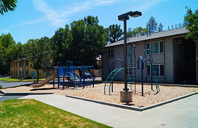 A photo of Brickyard Creek Apartments that shows the playground in the foreground. There is playground bark on the ground of the playground and green bars for children to climb on. In the background, is one of the buildings of the apartment complex.