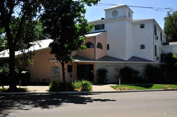 The photo is an exterior of the Campbel Commons apartment complex showing the front of the building. In the foreground is the road, then the building rises up showing its adobe color and white color exterior walls.