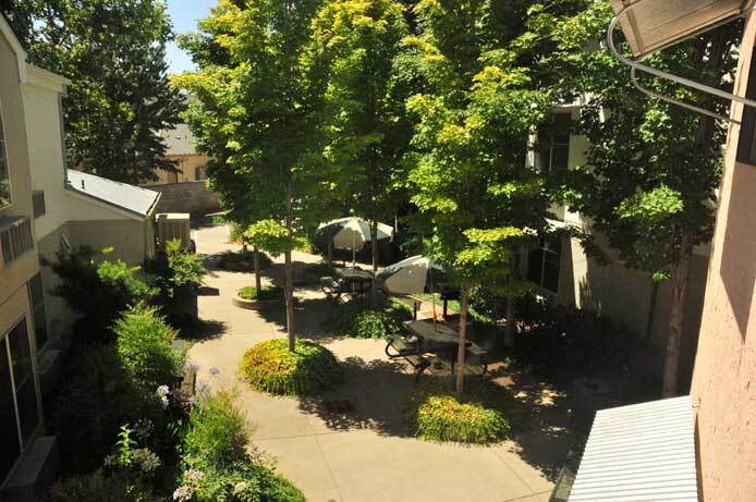 A photo taken from the second story shows the patio below, a mix of concrete and landscaped islands. Two tables with umbrellas are positioned in the shade of trees.