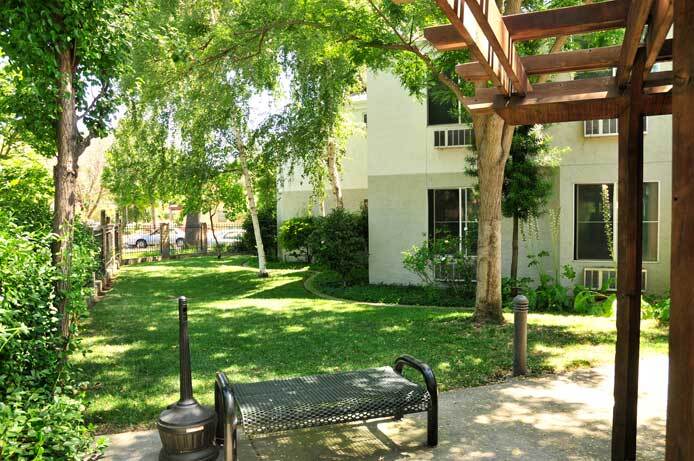 The photo is the exterior of the patio area. A grassy area shaded by trees is next to a shade structure. A bench is in the foreground.