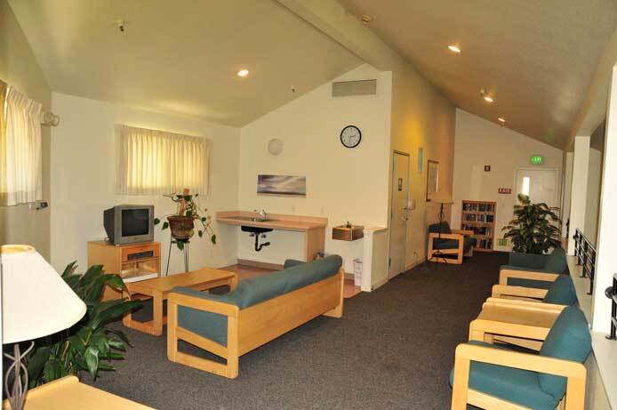 The photo shows one of the upstairs community areas. A sofa is next to a TV. Chairs line one side of the wall. There is a clock on the wall.