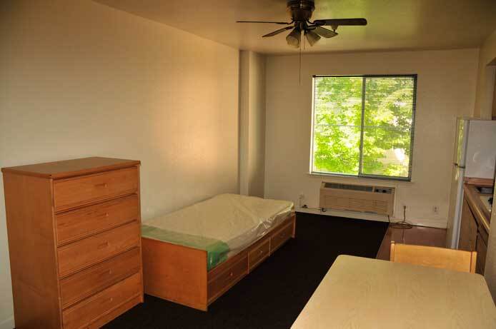 A photo of the studio apartment shows a dresser and a bed. There is a window and beneath it an AC unit. Along one side of the wall is a refrigerator.