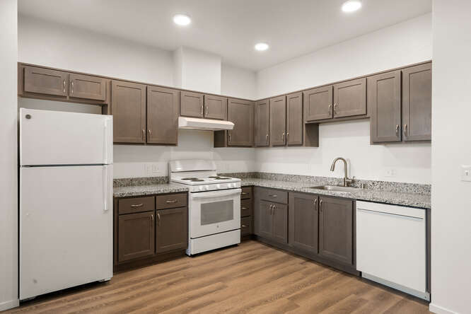This is an interior photo showing the full kitchen. The floor looks like a wooden floor. The cabinets are brown. The countertops look like a stone surface and are speckled white and gray. The 3 appliances are all white: Fridge, stove/oven, and dishwasher. The sink looks like stainless steel. There are three recessed lights in the ceiling above the kitchen.