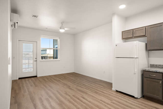 This is an interior photo showing the living room and partially showing the kitchen. The floors look like wooden floors. The walls are white. The door has glass and leads to the balcony from the living room. There is a window next to the door.  On the ceiling above the living room is a ceiling fan with an integrated light and three fan blades. The open area leads to the kitchen. Brown cabinets frame a white fridge. The fridge has a freezer on the top section of the fridge.