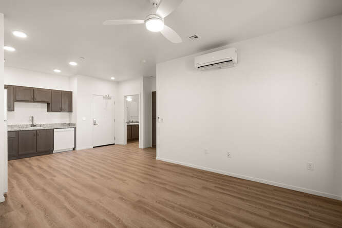 This is an interior photo showing the living room and partially showing the kitchen. The flooring looks like a wooden floor and the walls are white. In the center of the living room is a ceiling fan with an integrated light. The fan has 3 fan blades. One wall of the living room has a modular AC unit that is near the ceiling. The open area leads to the kitchen where this are brown cupboards a sink and a dishwasher. The remaining kitchen area is outside of the view.