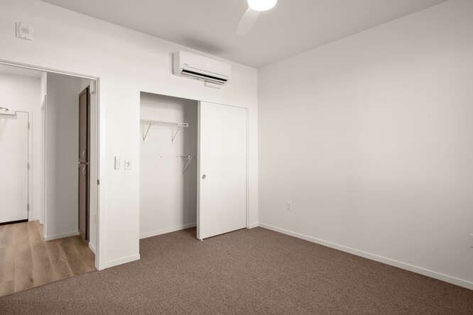 This is an interior photo showing the bedroom of one of the units. The floor has brown carpet, and the walls and ceiling are white. One wall has a door and a closet. The closet door is open. Above the closet is a modular AC unit. The wall next to it is a long wall with nothing on it. The walls are white.