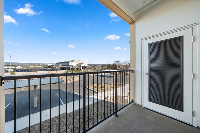 This is a photo taken on the balcony of a second-story unit. The photo shows a door on the side of the balcony, a corner of the balcony's floor, and the length of the metal railing. The view from the balcony shows the front cover parking lot and further in the background is Marsh Junior High School. The sky is bright blue with a few scattered clouds.