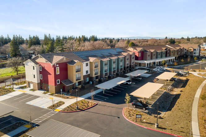 This is an exterior, aerial photo that shows the full length of the apartment building plus the covered parking lot in the front. The exterior of the building has multiple colors: red, yellow, green, and soft pink rise vertically on the 3-story building.