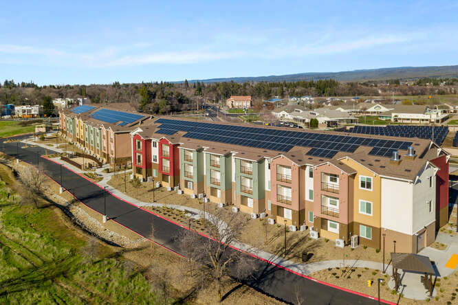 This is an exterior, aerial photo of the entire Creekside Place Apartments. The angle shows the opposite side from the front of the complex. There is a long asphalt road that runs along this area. A concrete sidewalk weaves along the road. The apartment building has solar panels along most of its roof. The exterior of the apartment is multiple colors, orange, soft pink, green, and red that rise in vertical columns of the 3-story building. In the background, there are indistinct buildings and many trees. The sky is light blue with a few soft clouds stretching horizontally.
