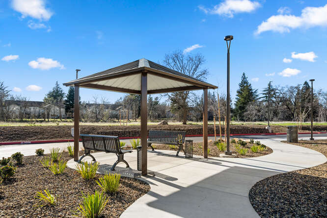 An exterior photo of a covered ten-foot by ten-foot patio. A concrete pathway curves through and connects to the patio. Around the patio is landscaping, brown gravel with green shrubs scattered. Under the patio covering are two benches. A tall light pole stands off the to the side of the patio and next to the pathway. In the background, there are trees and a bright blue sky with some scattered clouds.