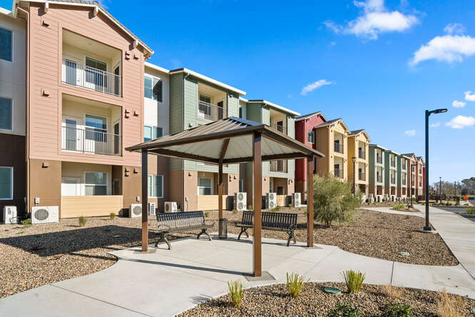 An exterior photo of Creekside Place Apartments shows a three-story apartment complex on a sunny day with just a few clouds in a bright blue sky. In the foreground is a ten-foot by ten-foot covered patio with two benches. A concrete walkway connects to the patio and runs the length of the long apartment complex. The apartment building has a mix of colors rising vertically: soft pink, green, red, and yellow blocks of color. The covered balconies frame glass doors and windows of the apartment units.