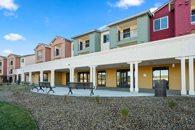 An exterior photo of Creekside Place Apartments showing the front landscaping and patio. Brown gravel and shrubs make up the landscaping and the concrete patio is a semi-circle with two benches and a trash can. Above the patio is a white awning the spaces the length of the long apartment building. Underneath the awning is more patio space, empty, that then meets the exterior wall with 4 large glass doors that open up to the patio. The second story of the apartments features multi-color exterirors: Red, green and a soft pink scattered between white areas. The balconies of the second story units are there. They are covered balconies with glass doors and windows.