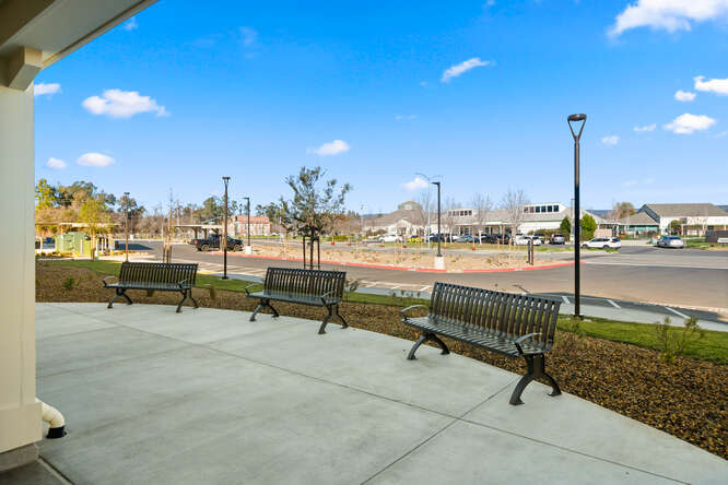 An exterior photo of Creekside Place Apartments taken from the vantage point underneath the front patio. The ground of the patio is a semi-circle of concrete with 3 mental benches facing the front of the apartments. Behind the patio, a section of landscaping with gravel, and shrubs meets a section of grass where two narrow and tall light poles rise into the blue sky with a few scattered clouds. Beyond that area is a sidewalk and the front driveway of the apartment complex. Further in the background is Marsh Junior High.