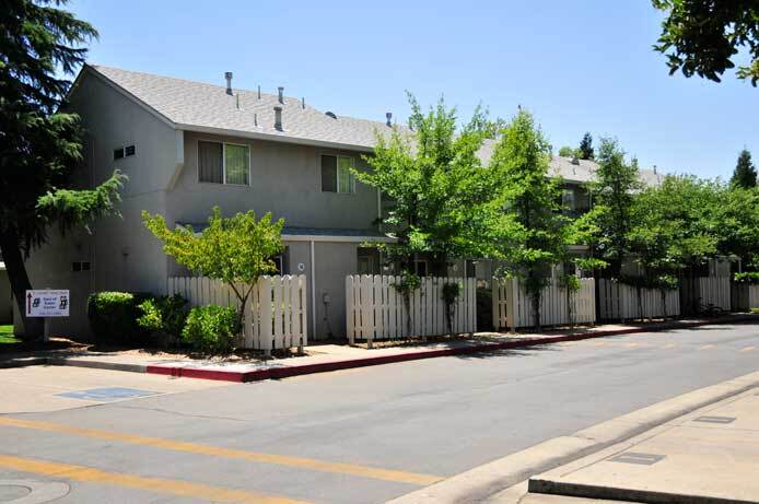 An exterior photo of the apartment building shows a two-story building. The asphalt road runs next to the buildings where there are small fenced patio areas. Several trees line the road.