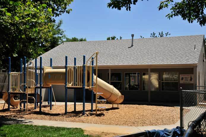An exterior photo shows a playground. The playground has a slide on one end and another smaller slide on the other end. Between the two slides are platforms that you can walk on. The ground beneath the playground is covered in playground bark. In the background of the photo, near the playground is the community room.